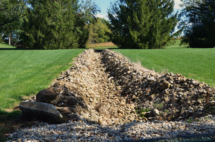 An open drainage trench filled with stone running across a mown lawn between two areas of grass